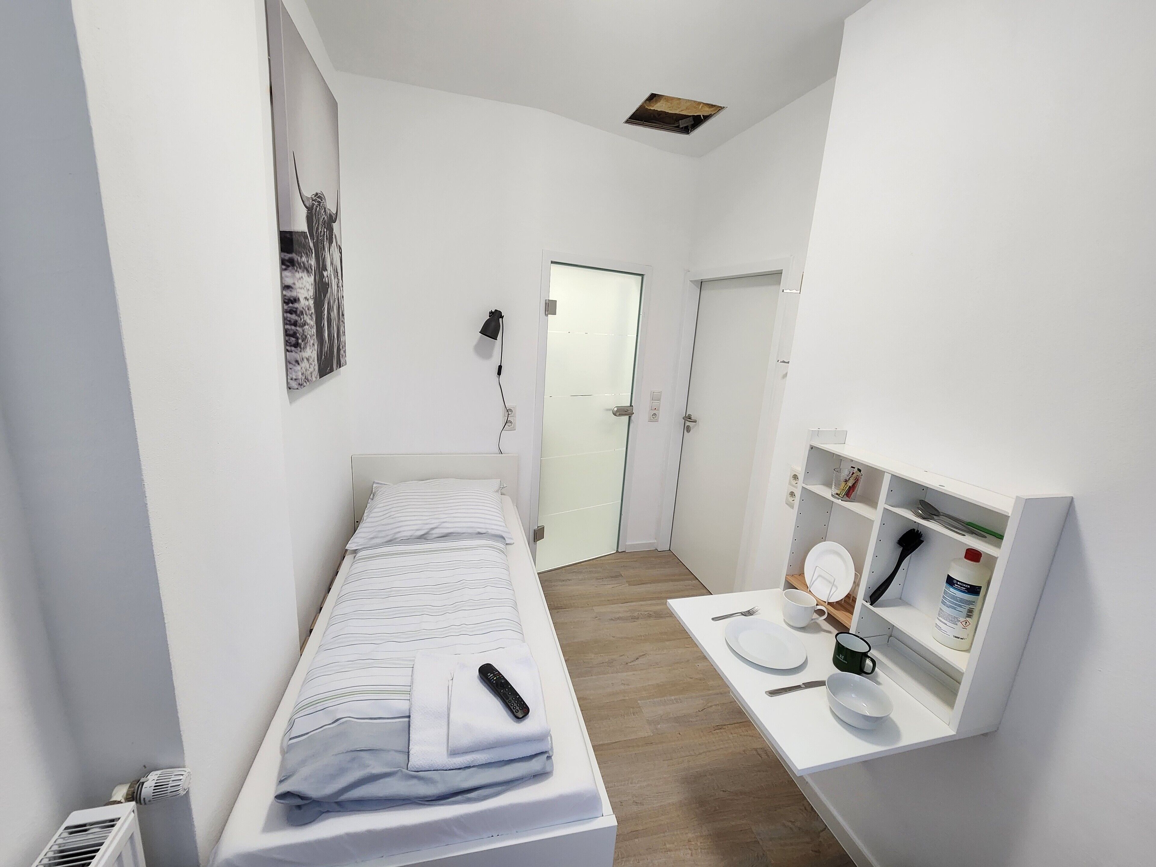 A small, narrow hotel room with a single bed, a bedside table with a remote control, a small shelf with toiletries, and a door leading to another room.