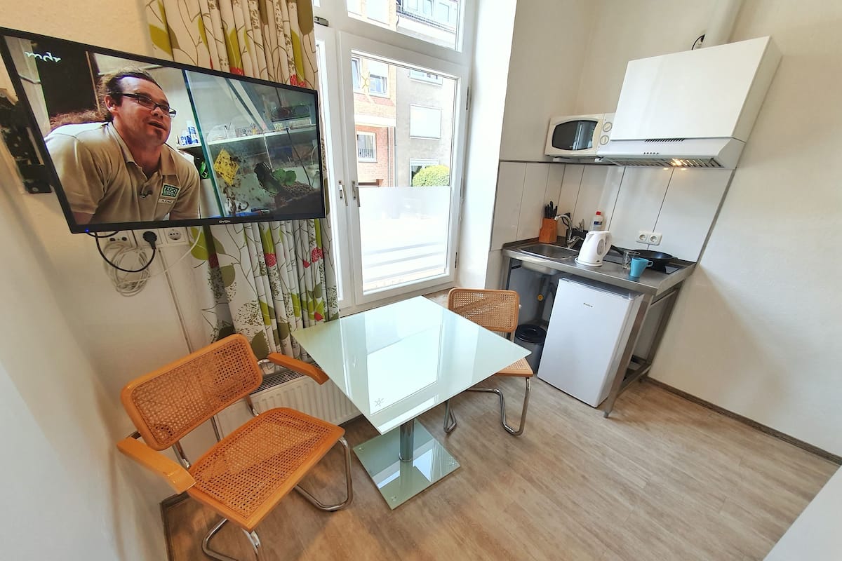 A compact kitchen with a dining area, a television mounted on the wall, and a window with curtains.