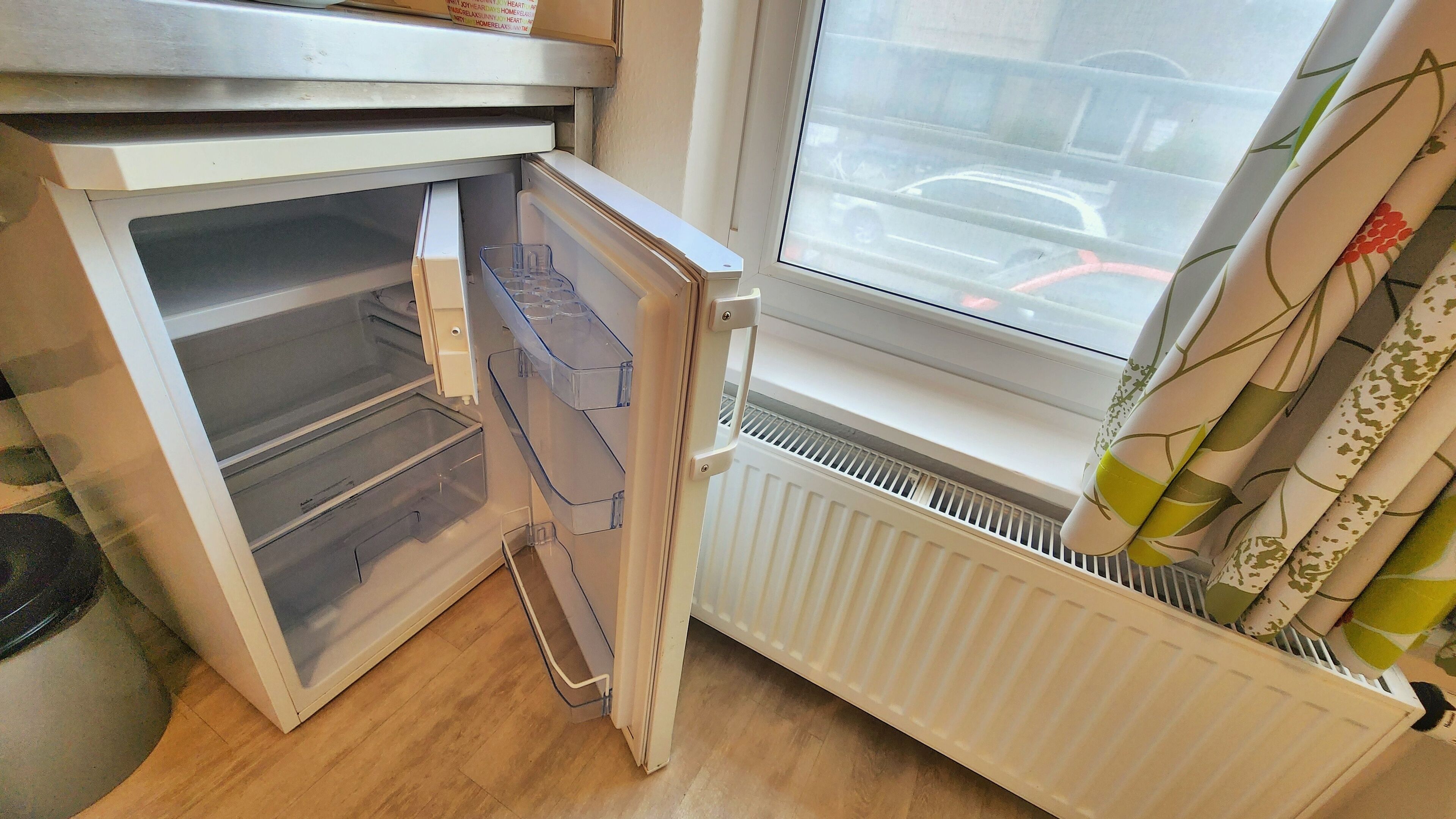 Ein offener Kühlschrank mit durchsichtigen Kunststoffbehältern im Inneren, ein Fenster mit Blick auf einen Parkplatz und ein Heizkörper unterhalb des Fensters.
