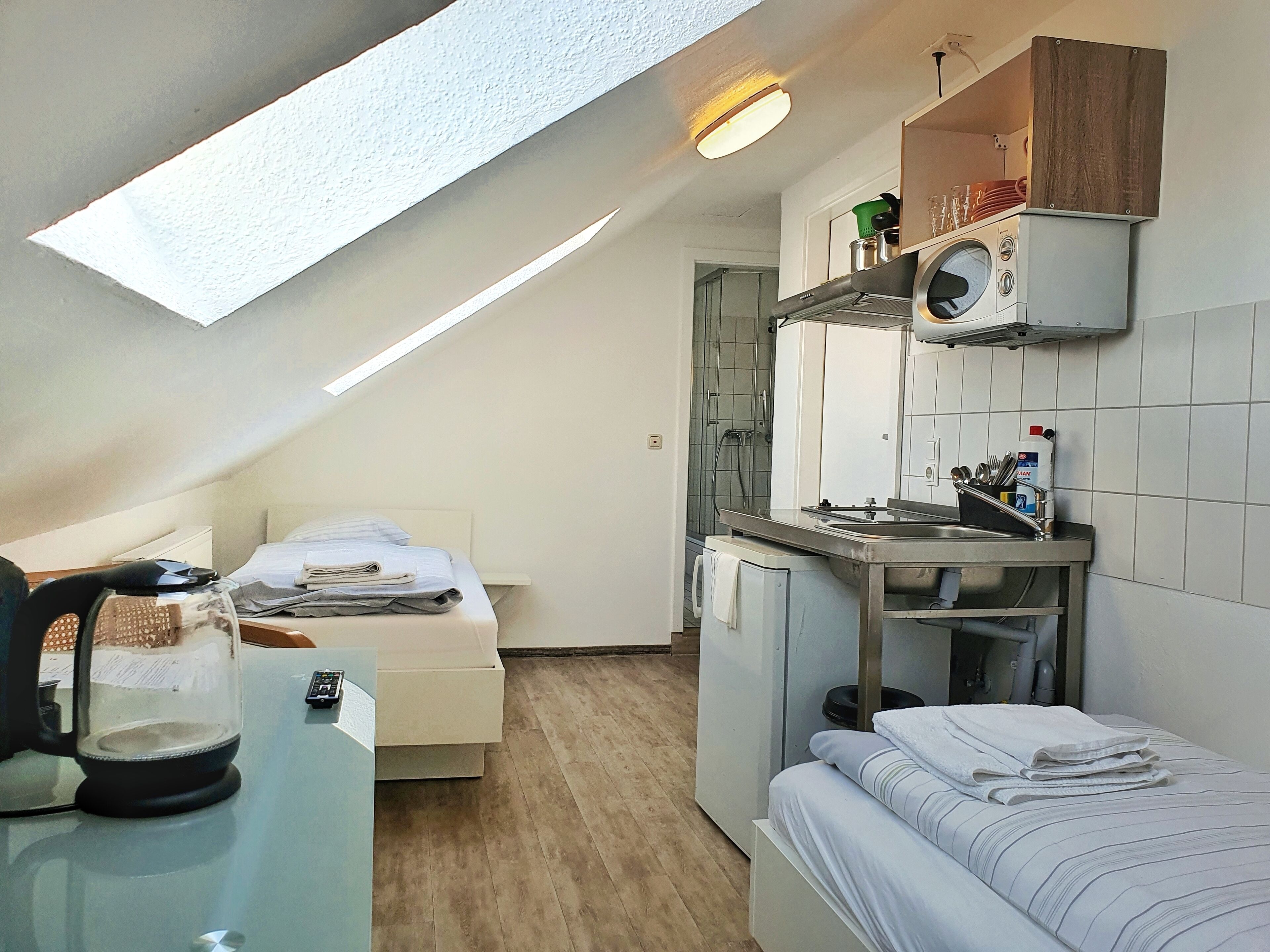 Ein kleines Zimmer mit Bett, Schreibtisch mit Wasserkocher, einer kleinen Küchenzeile mit Mikrowelle und einem Oberlicht.