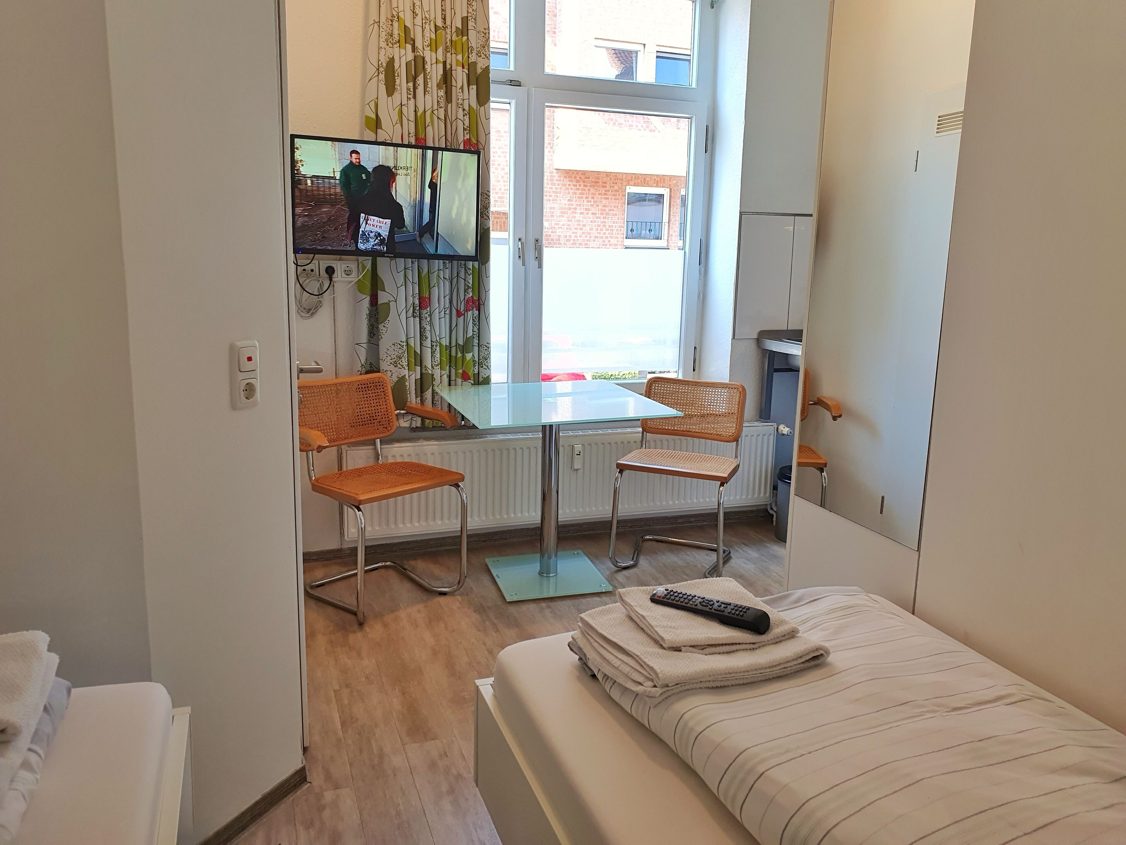 A small, well-lit room with a bed, two wicker chairs, a table, and a TV mounted on the wall.
