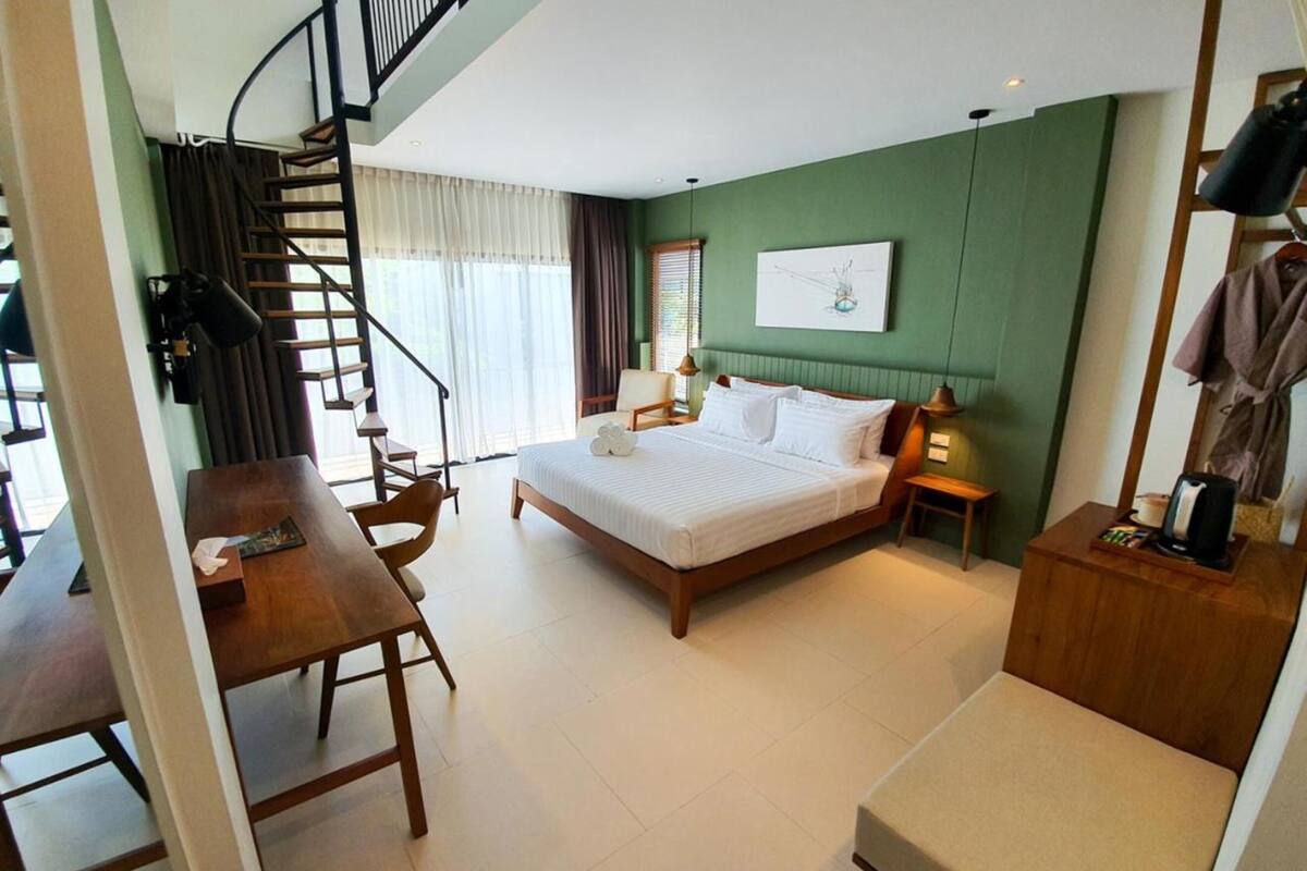 A bedroom with a large bed, a wooden desk, a chair, a small table, a lamp, and a painting on the wall.