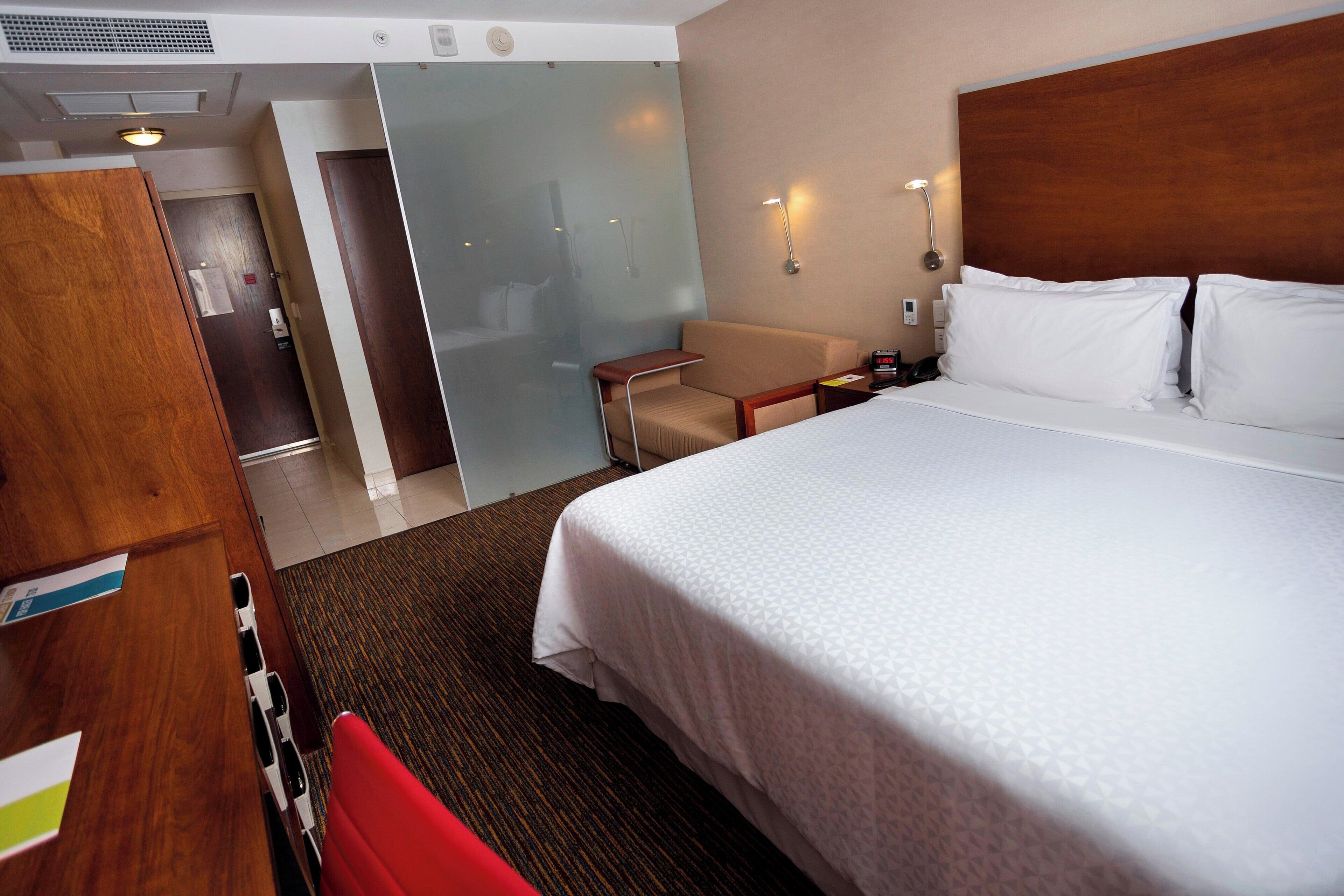 A hotel room with a bed, a desk, a chair, a small table, and a wall-mounted lamp.