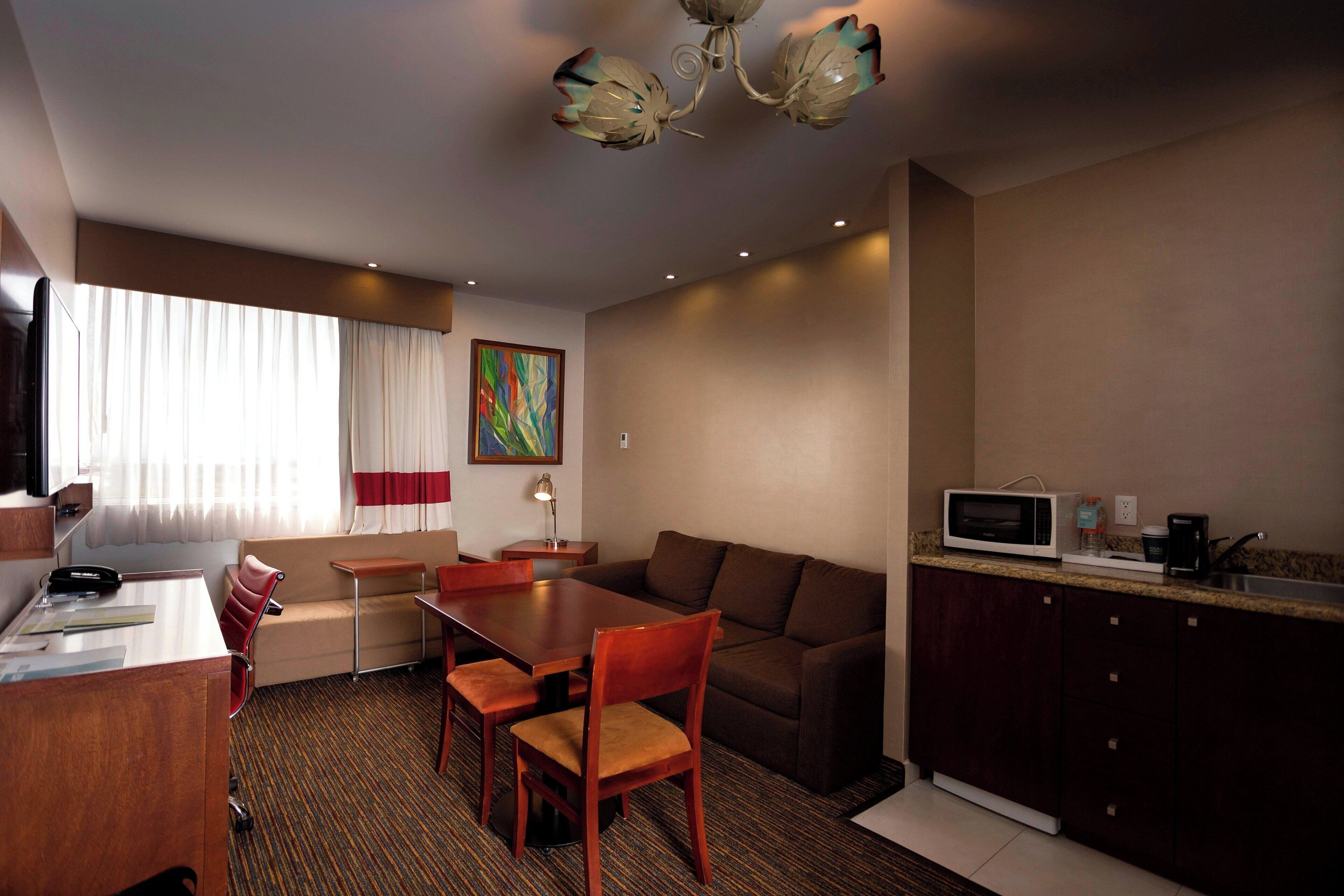 Habitación de hotel con mesa de comedor, sillas, sofá, televisor, microondas y pequeña cocina.