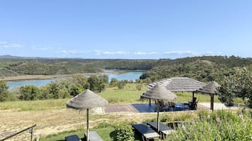 Un espace extérieur paisible, agrémenté de parasols en chaume, de chaises longues et offrant une vue sur un lac et des collines boisées.