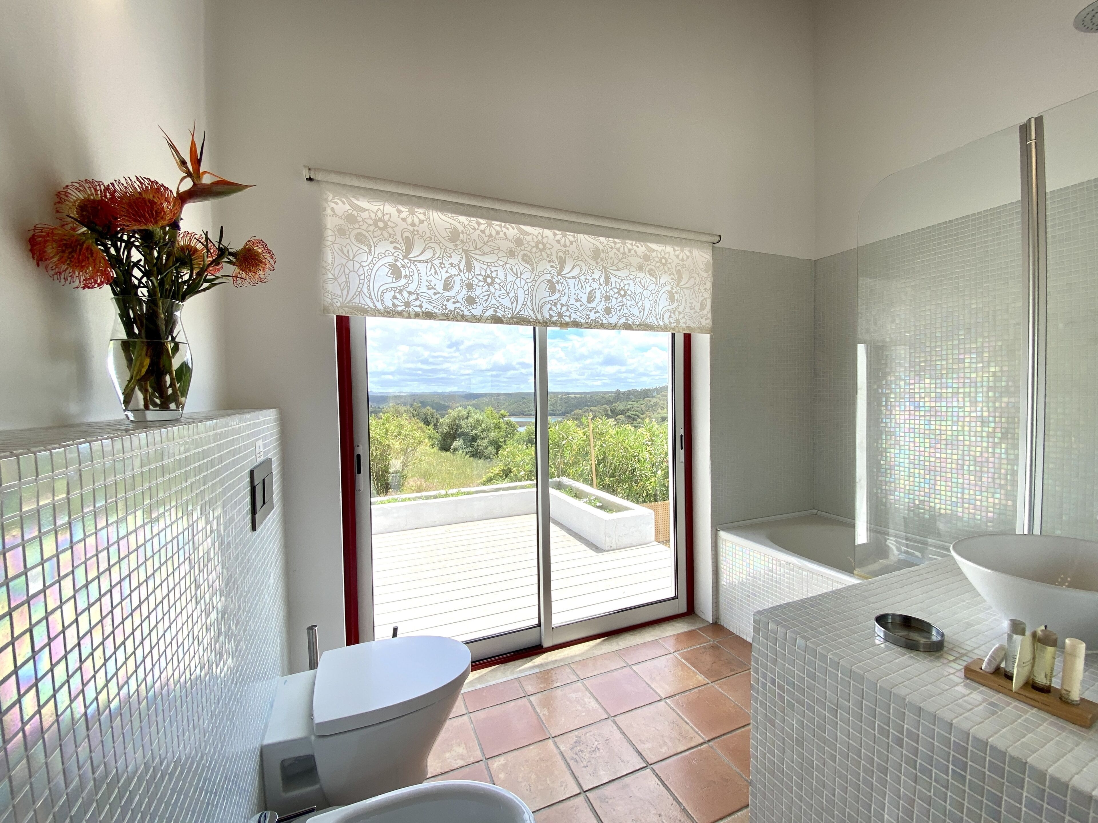 Une salle de bain équipée de toilettes, d’un bidet et d’un lavabo, un vase avec des fleurs et une vue à travers une porte coulissante.