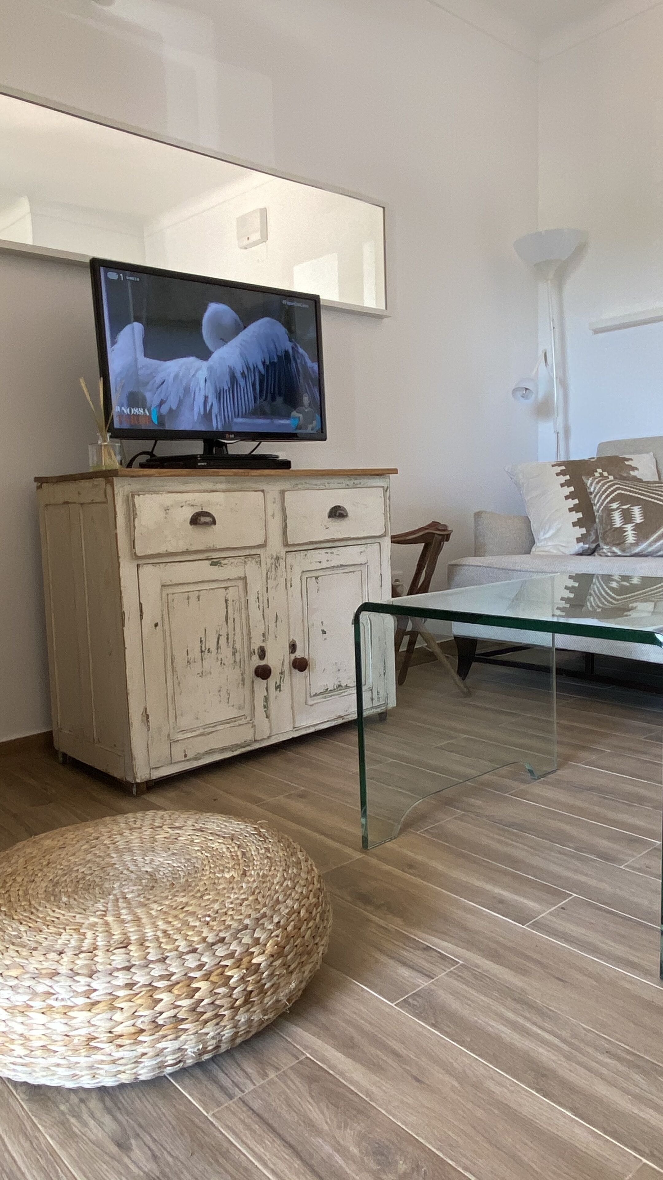 A modern living room with a television on a white cabinet, a glass coffee table, a woven ottoman, and a sofa with patterned cushions.