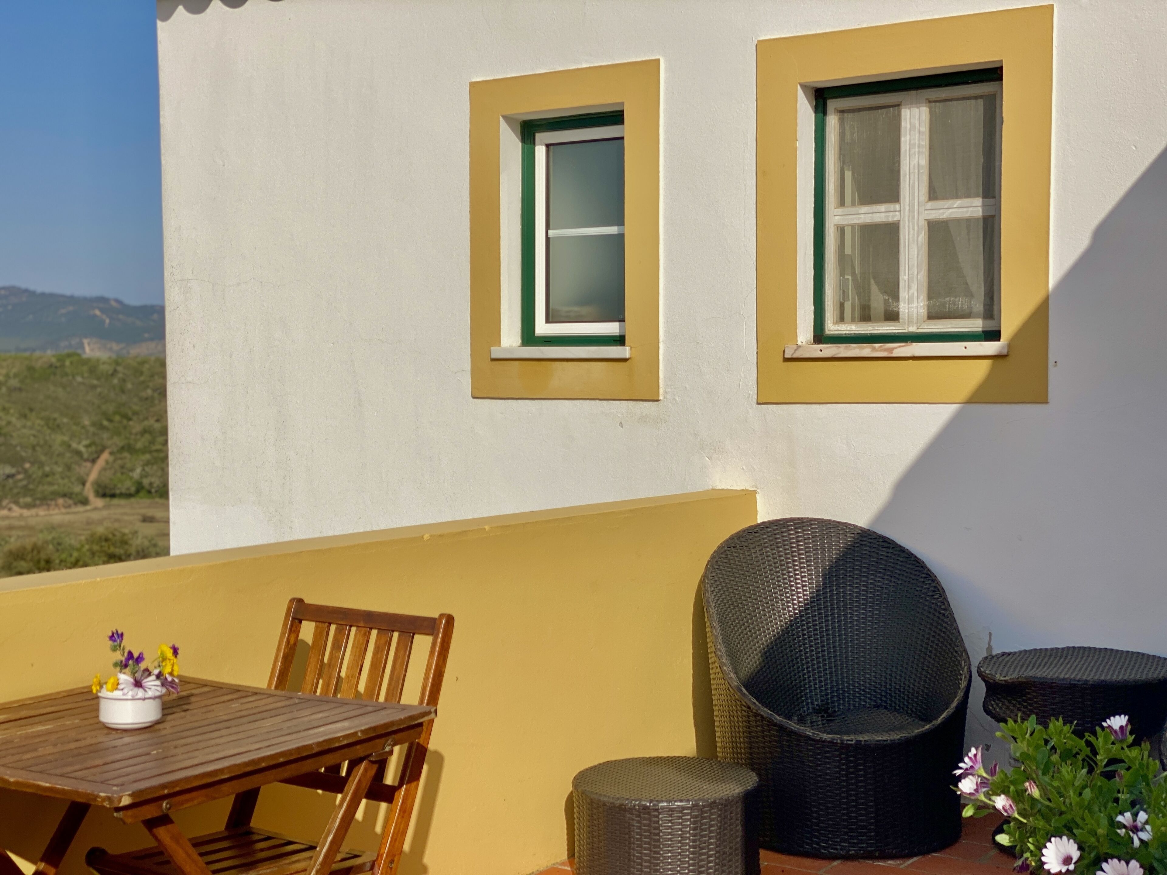 Uma varanda com mesa e cadeiras, duas janelas com molduras verdes, e vista para o campo.