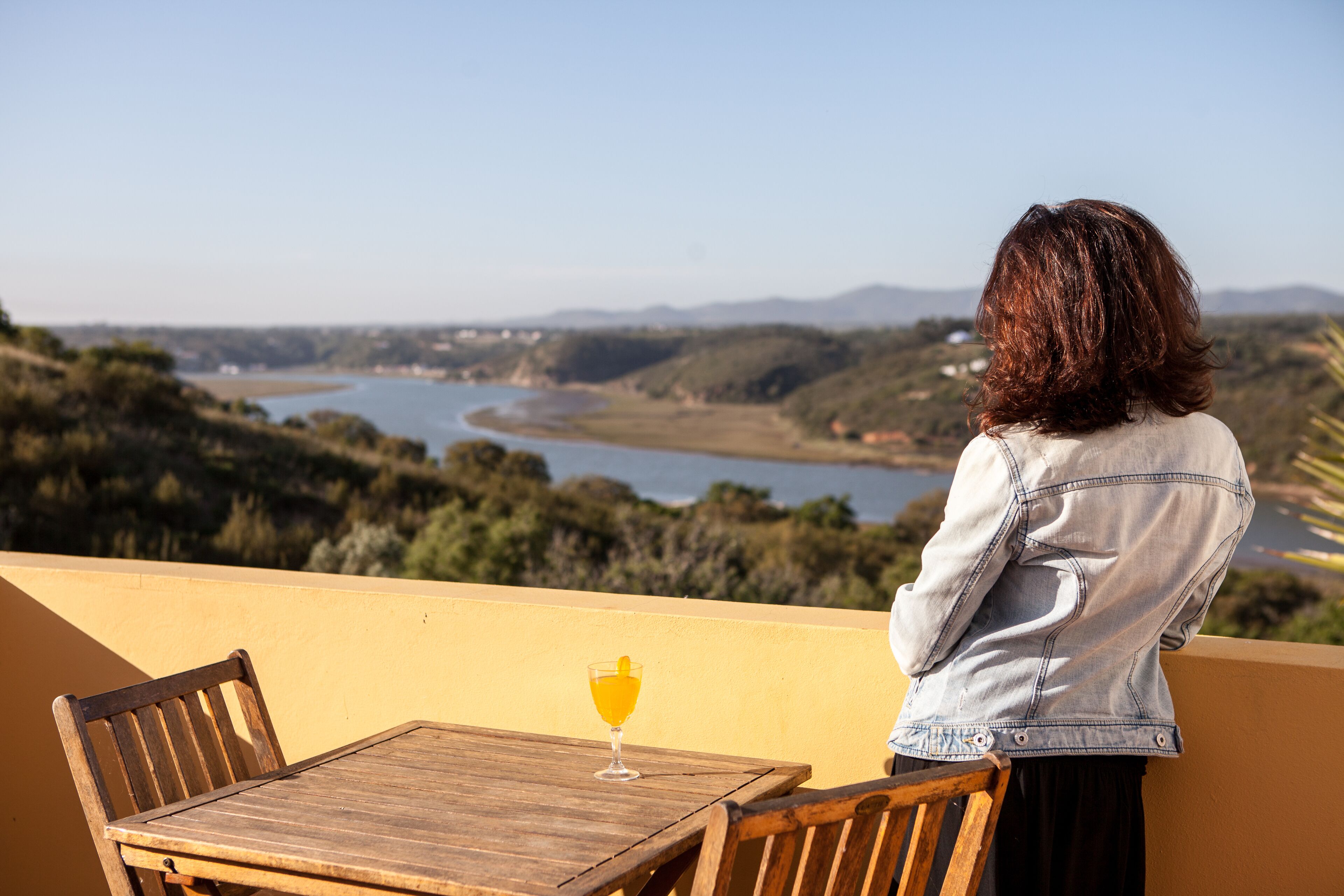 Una persona sentada en una mesa de madera, con un vaso de jugo de naranja, contemplando un paisaje con colinas y un río.