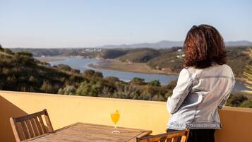 Una persona sentada en una mesa de madera, con un vaso de jugo de naranja, contemplando un paisaje con colinas y un río.