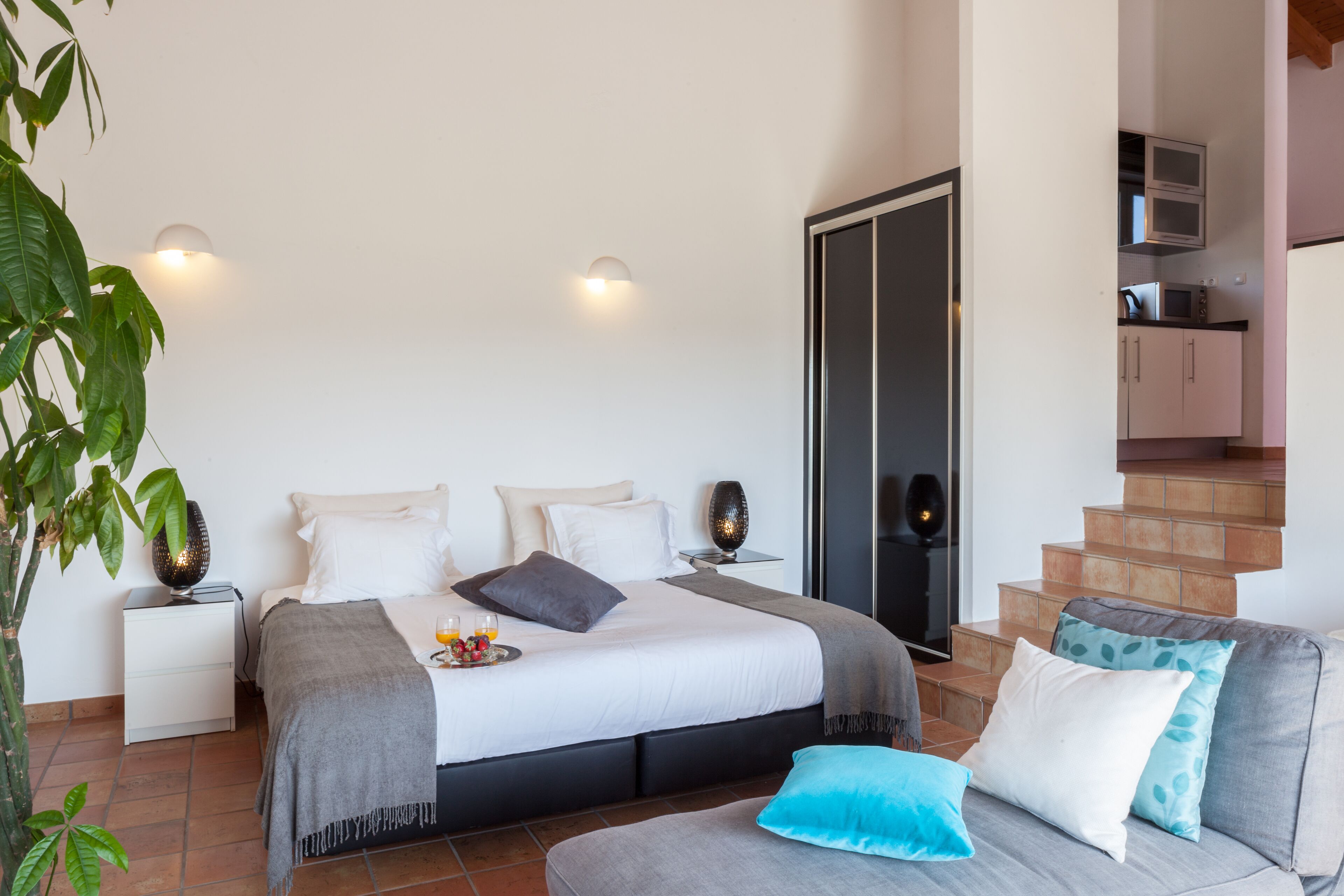 Quarto com cama, mesas de cabeceira, um criado-mudo, televisão, escada e área de cozinha.