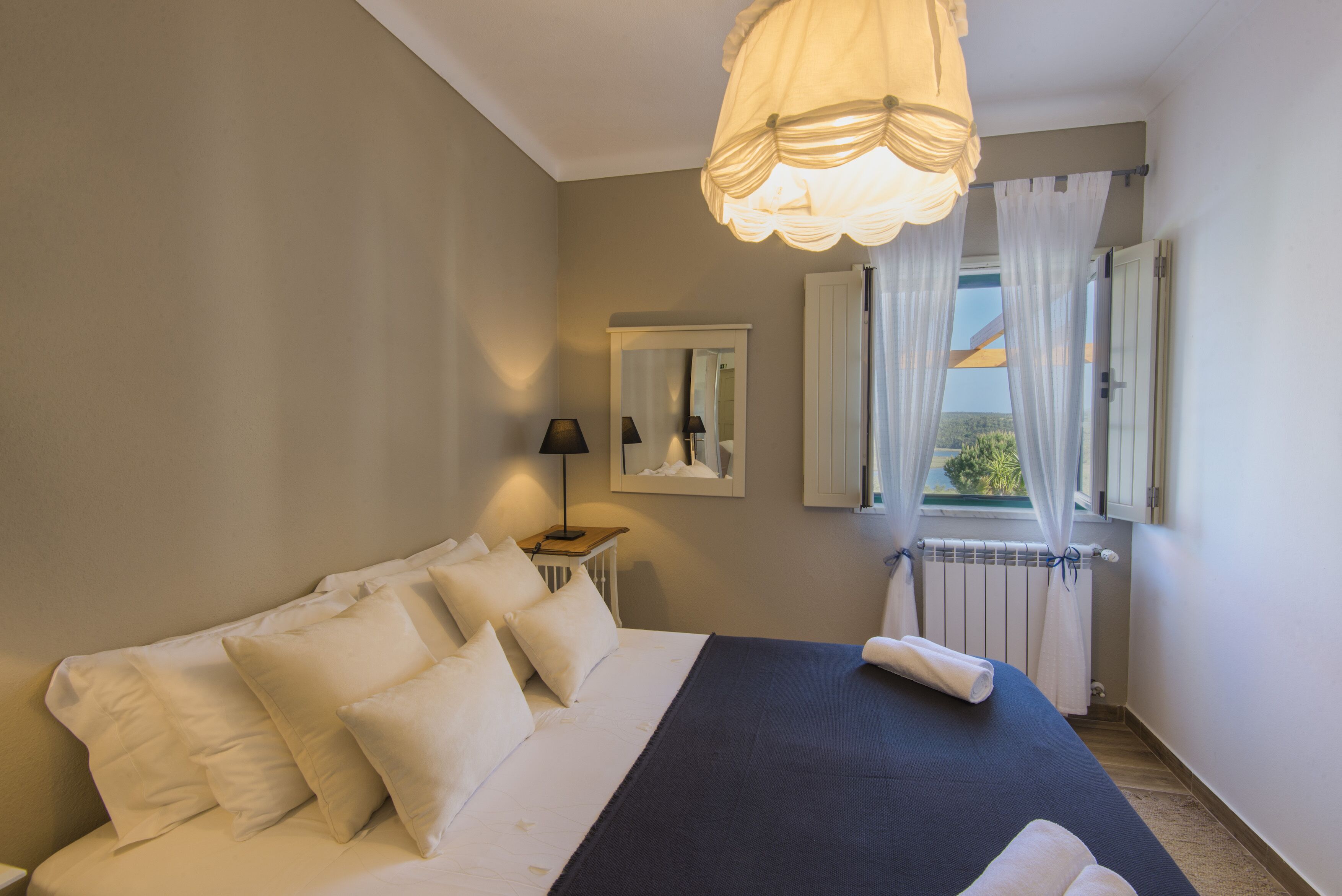 Une chambre avec un grand lit, une table de chevet avec une lampe, une fenêtre avec des rideaux et une vue sur l’extérieur.