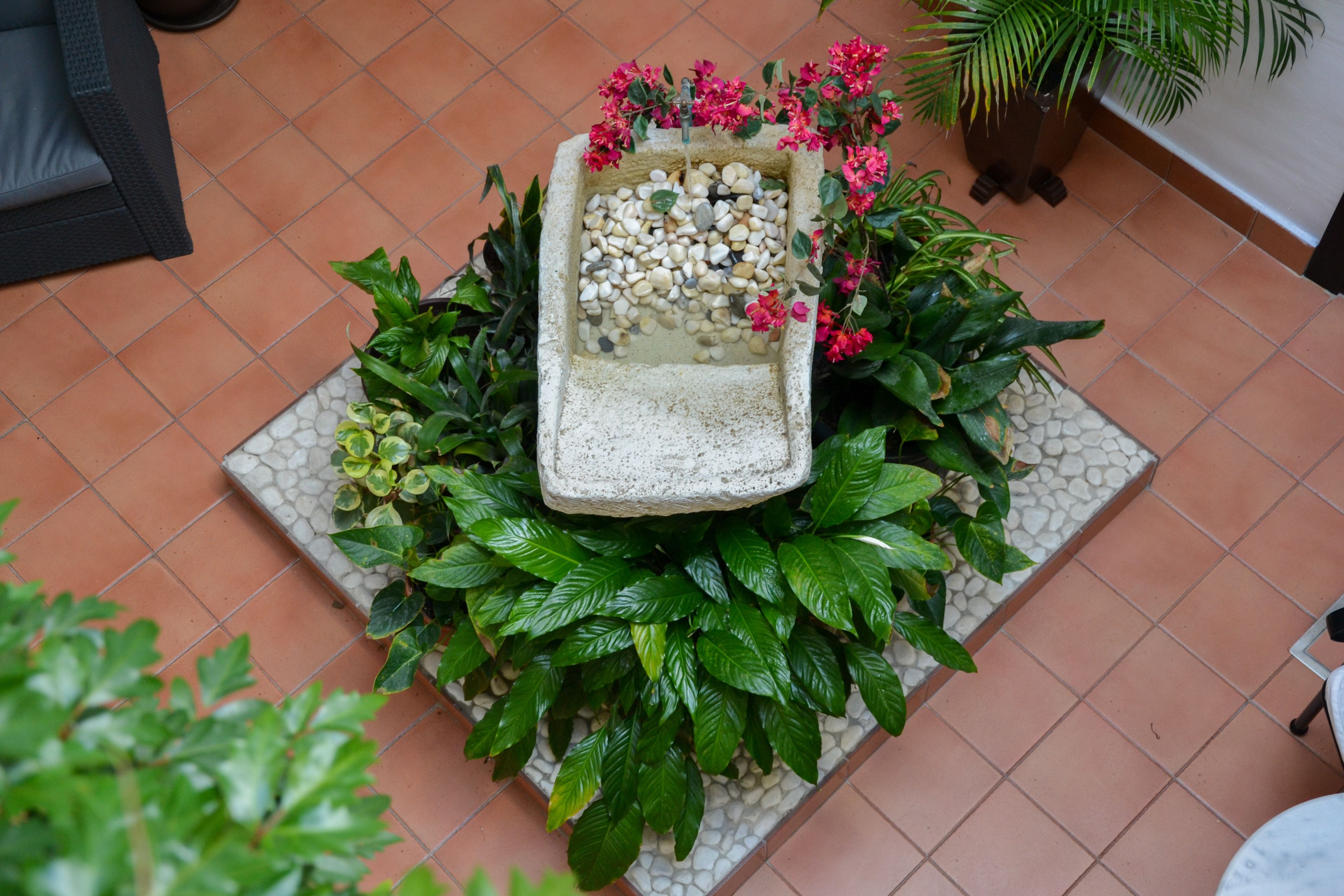 A small garden area with a stone planter filled with pebbles and pink flowers, surrounded by lush green plants.