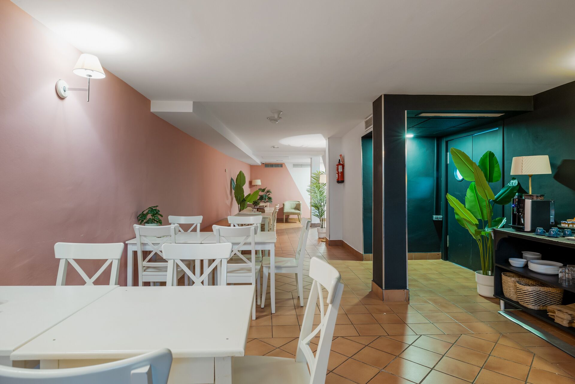 Un espace repas avec des tables et des chaises blanches, un coin cuisine avec un comptoir et des appareils électroménagers, et un mur végétal agrémenté d’une plante.
