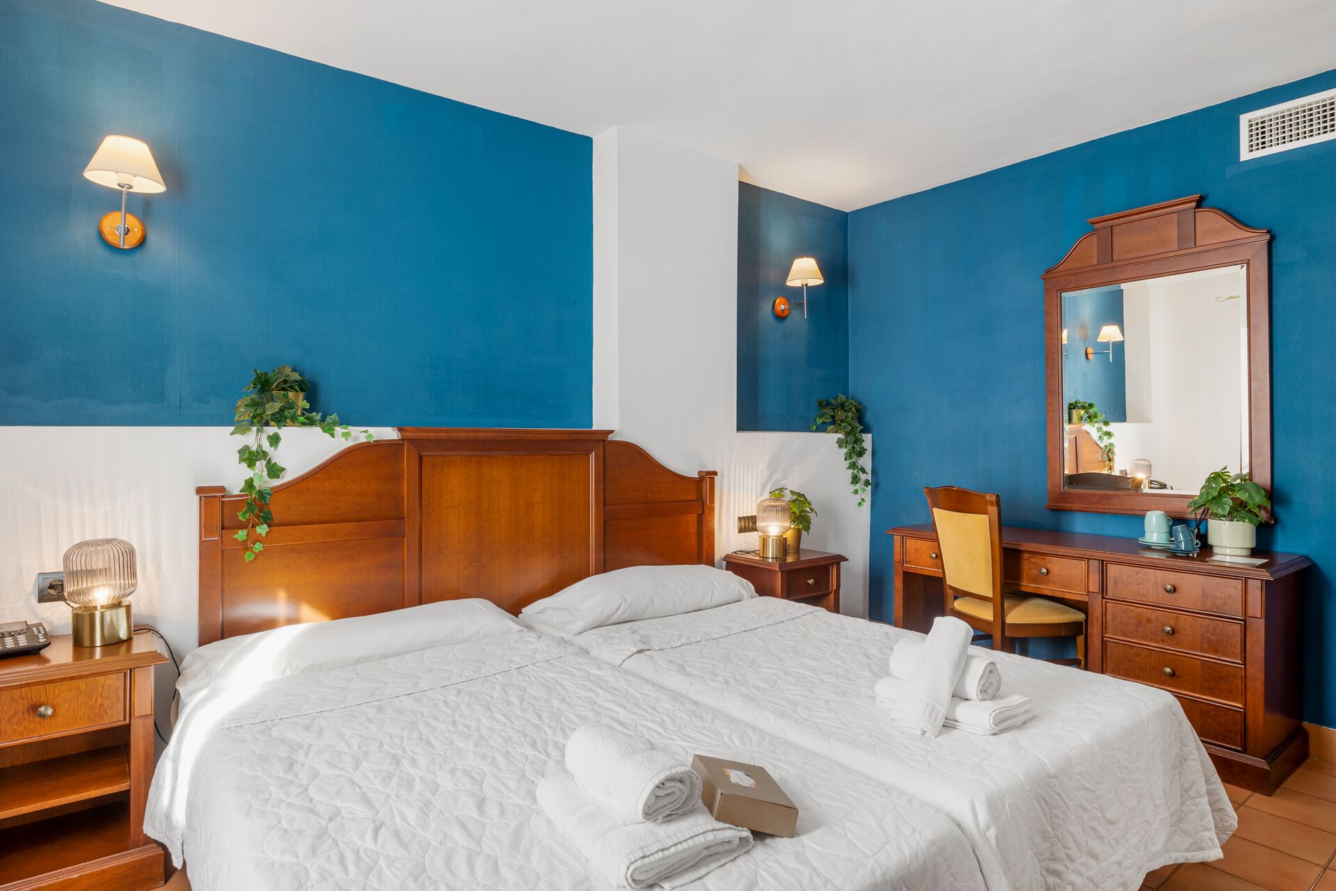 Habitación de hotel con una cama grande, cabecera de madera, dos mesitas de noche con lámparas, un aparador y un espejo.