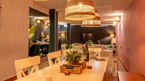 Un comedor con una mesa larga, sillas blancas, lámparas colgantes de mimbre y una planta en maceta.