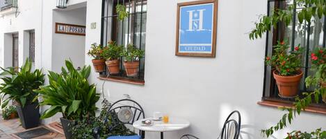 A small outdoor seating area with two metal chairs and a table, surrounded by potted plants and a sign reading "LA POSADA CIUDAD".