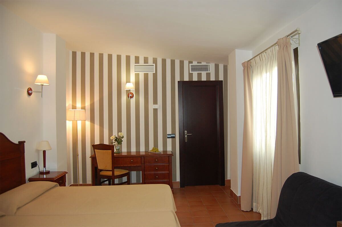 A hotel room with a bed, a desk with a chair, a lamp, a wall-mounted TV, and a door with curtains.