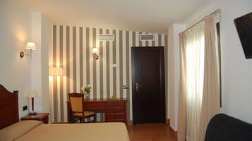 A hotel room with a bed, a desk with a chair, a lamp, a wall-mounted TV, and a door with curtains.
