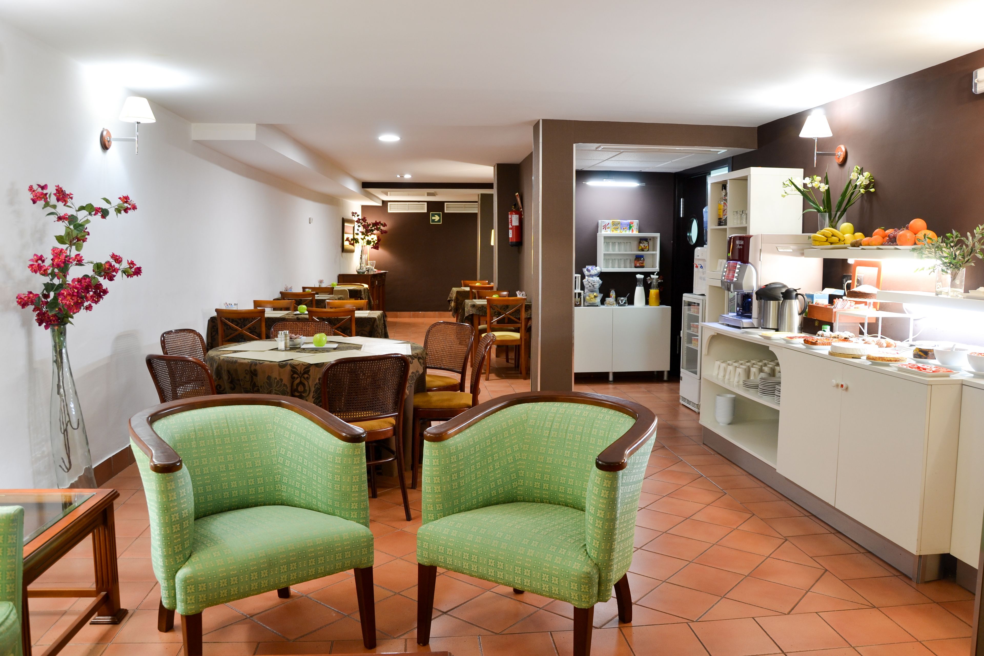 Comedor con sillas tapizadas en verde, una mesa redonda y una zona de cocina con cafetera y exposición de fruta.
