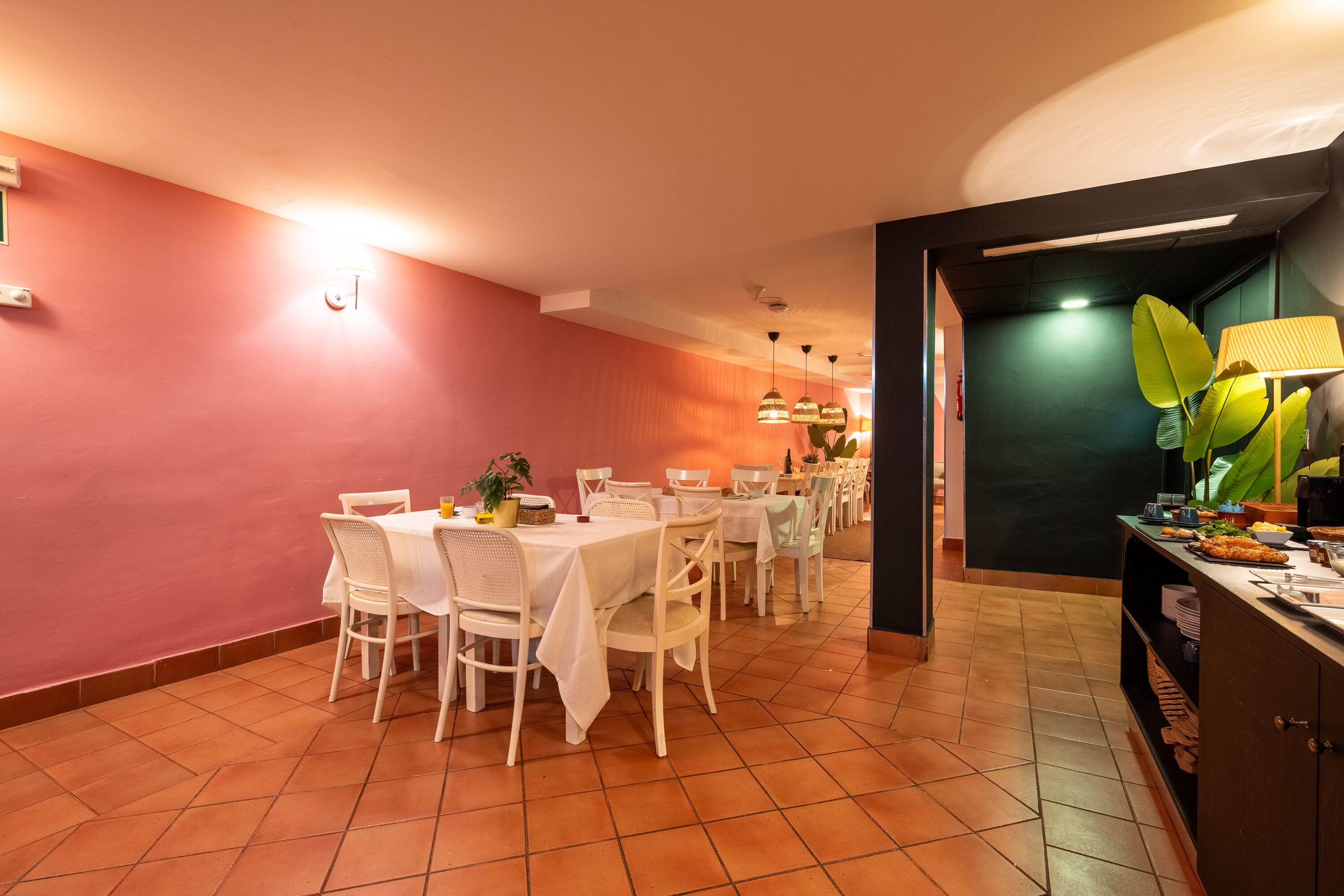 Un comedor con una mesa blanca dispuesta para seis personas, rodeada de sillas blancas, con una planta verde sobre la mesa y un mueble negro con una lámpara a la derecha.