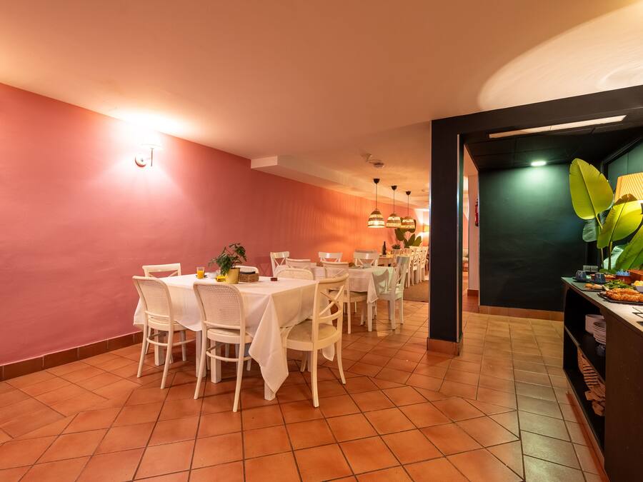 Un comedor con una mesa blanca dispuesta para seis personas, rodeada de sillas blancas, con una planta verde sobre la mesa y un mueble negro con una lámpara a la derecha.
