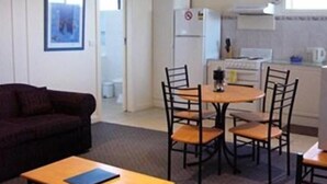 Un espacio habitable compacto con un sofá oscuro, una pequeña mesa de madera, una mesa redonda para comer con cuatro sillas, un refrigerador blanco y una zona de cocina con estufa y armarios.