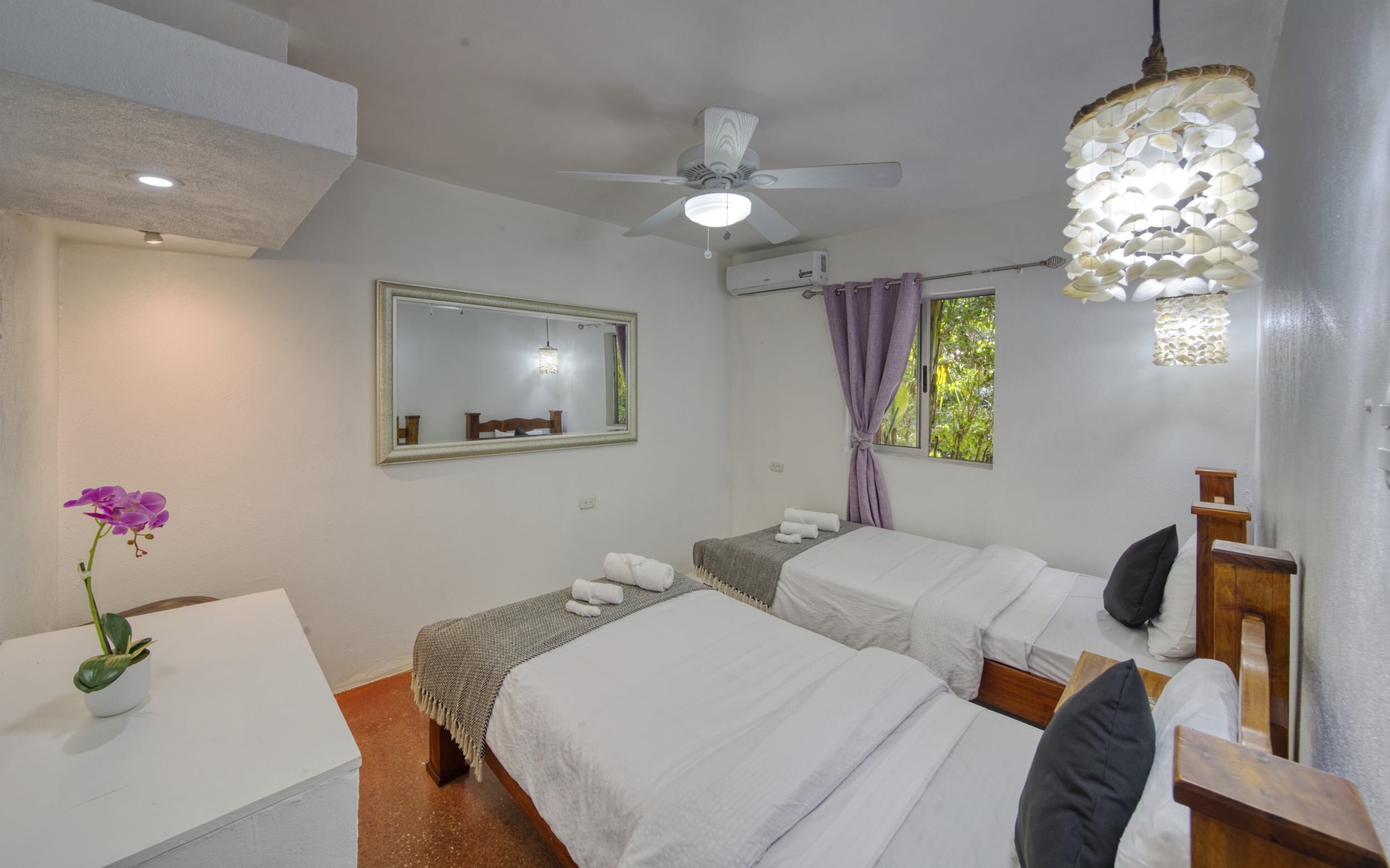 Une chambre d’hôtel avec deux lits, un ventilateur de plafond, un miroir, une fenêtre avec des rideaux et une petite table sur laquelle est posée une plante.