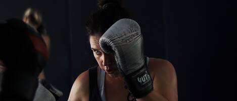 Una persona con camiseta oscura y pantalones cortos, usando guantes de boxeo, en una habitación tenuemente iluminada con fondo azul y un logotipo en la pared.
