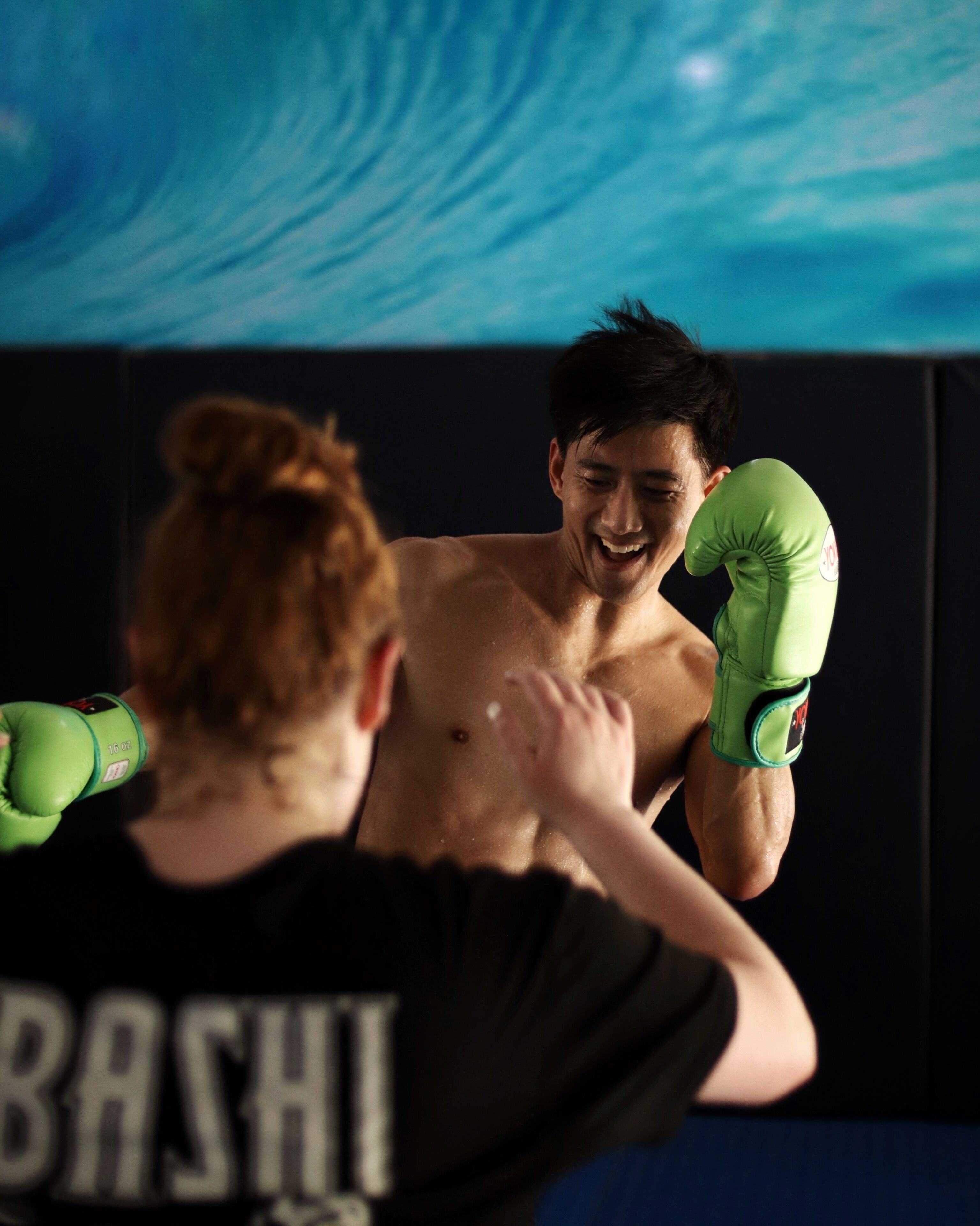 A person in a black shirt with "BASHI" on the back, practicing boxing with green gloves.