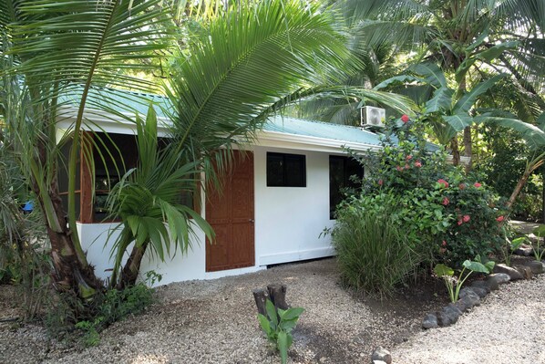 A small white building with a green roof, surrounded by lush greenery and a gravel path.