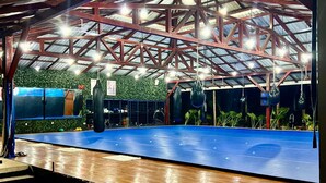 Een overdekte sportfaciliteit met een blauwe, verende vloer, metalen balken en hangende bokszakken.