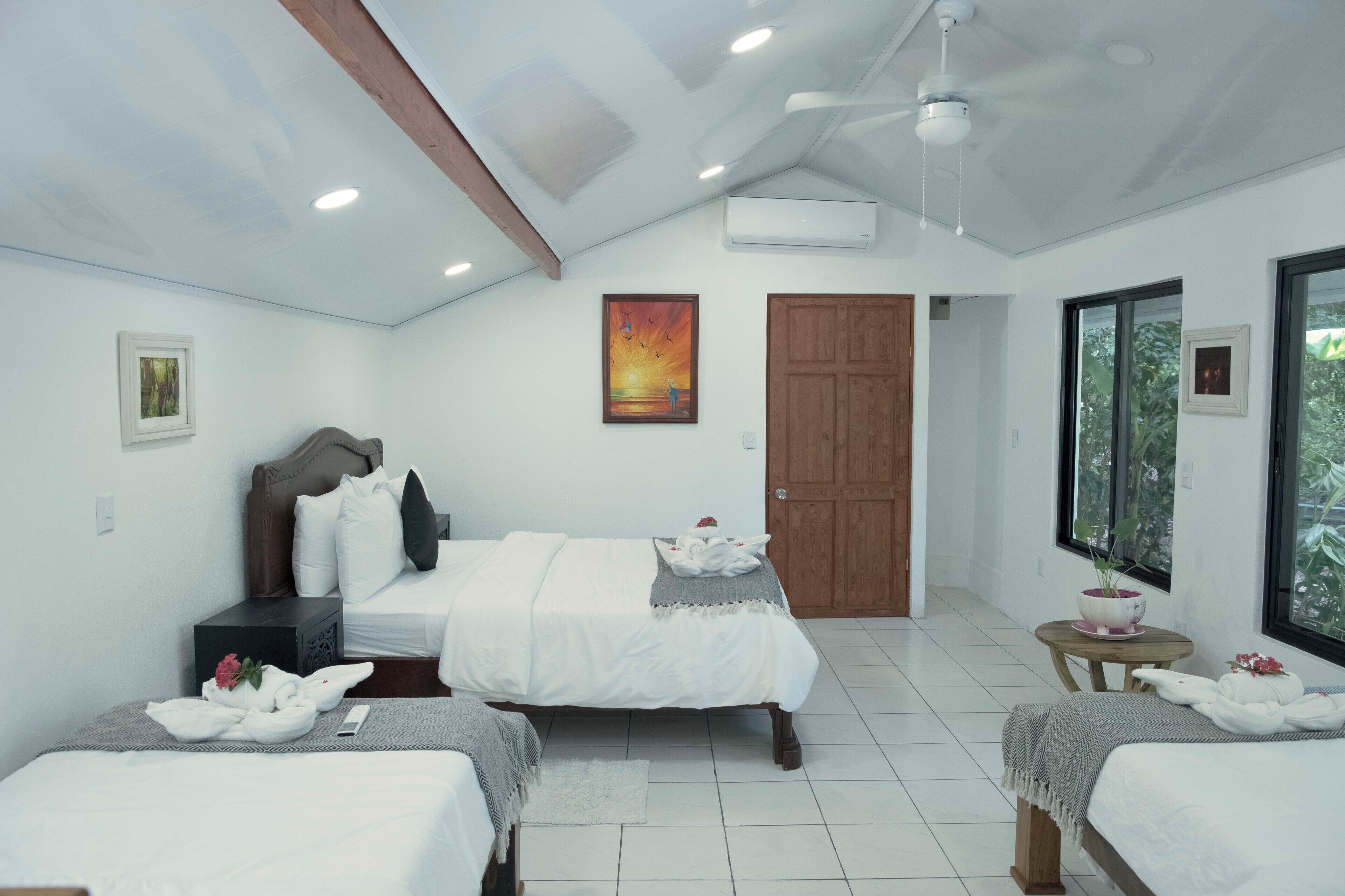 Une chambre d’hôtel avec deux lits, une tête de lit en bois, un ventilateur de plafond, un appareil de climatisation, un tableau et une fenêtre donnant sur un paysage verdoyant.