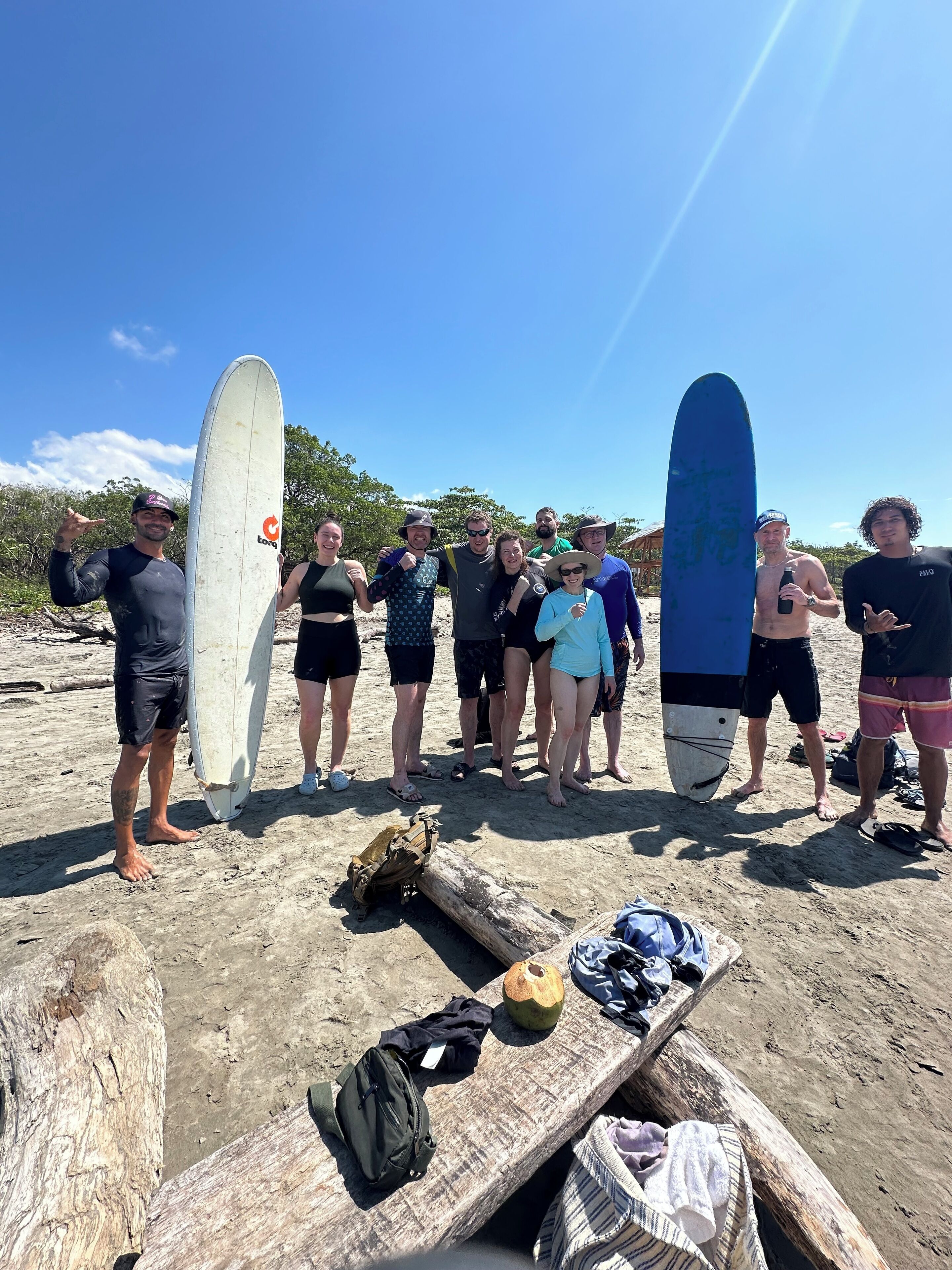 A group of people on a sandy beach holding surfboards, with a wooden bench and some bags in the foreground.