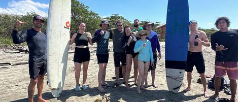 A group of people on a sandy beach holding surfboards, with a wooden bench and some bags in the foreground.