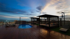 Une terrasse en bois avec une piscine, des chaises longues et un espace salon couvert offrant une vue sur la mer.