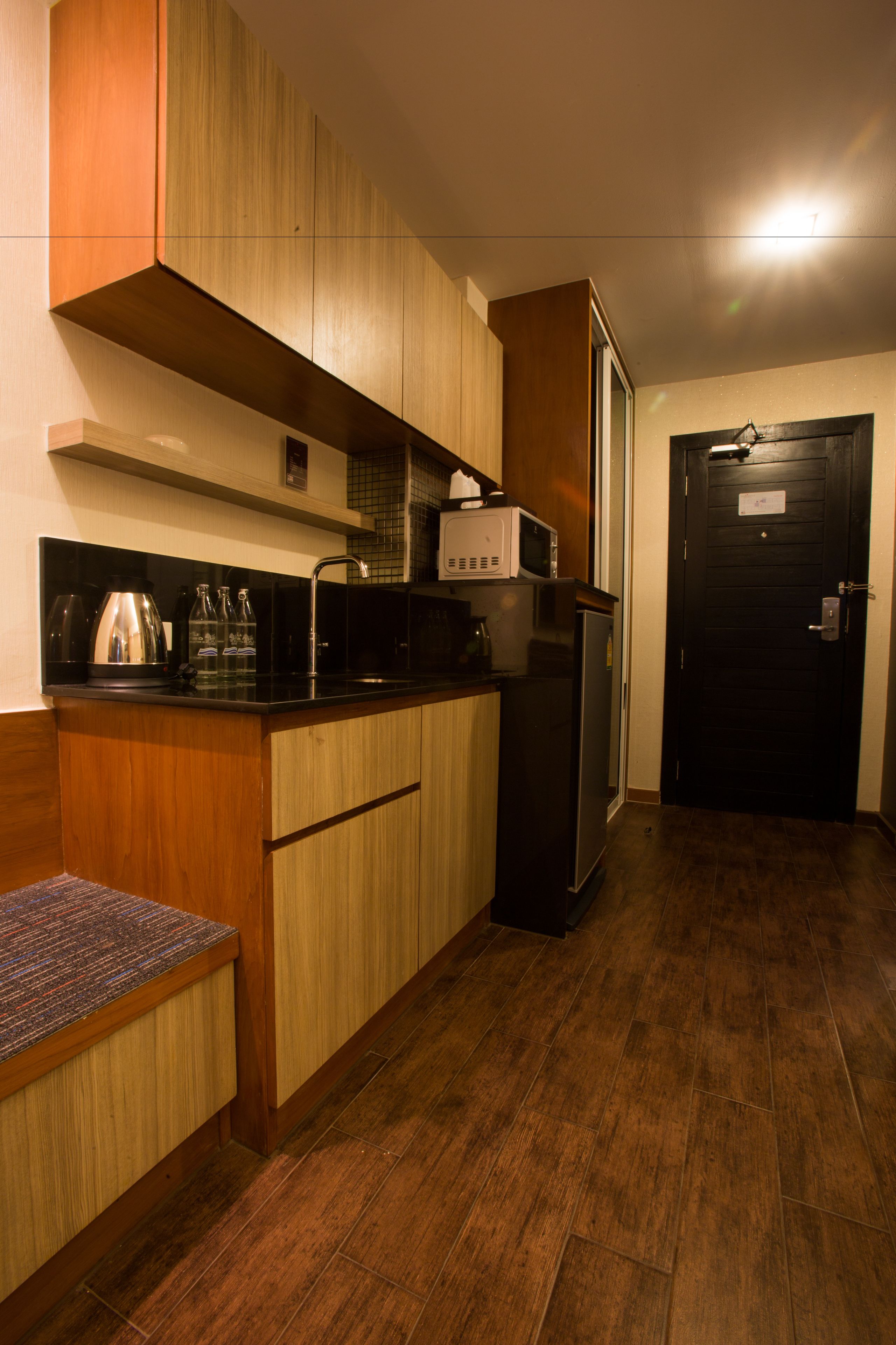 Cocina compacta con armarios de madera, encimera negra, microondas, refrigerador y un banco de madera.