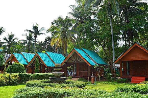 A row of wooden cabins with green roofs, surrounded by lush greenery and palm trees.