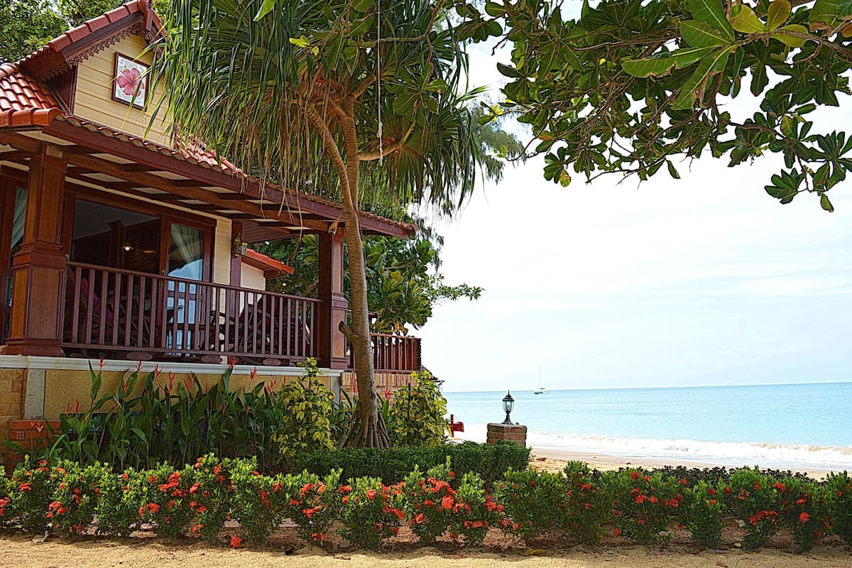 A beachfront building with a balcony overlooking the sea, surrounded by lush greenery and red flowers.