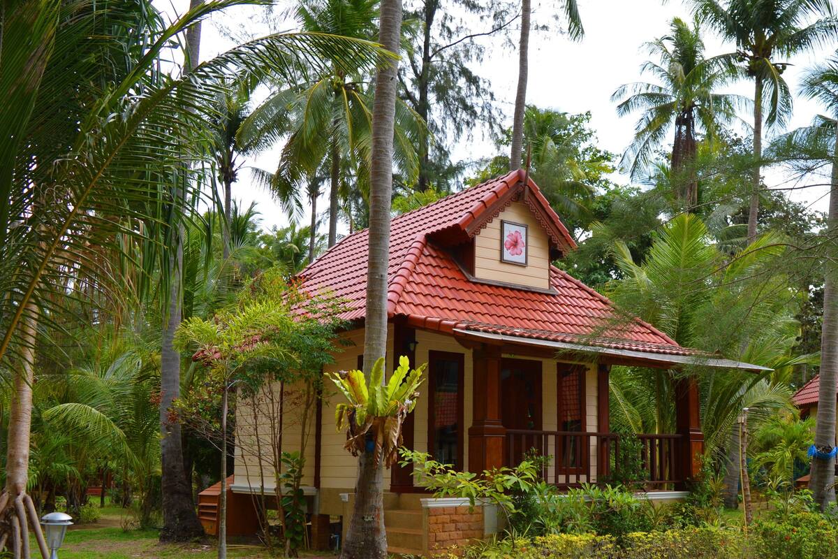 A small, single-story house with a red-tiled roof, surrounded by lush tropical vegetation.