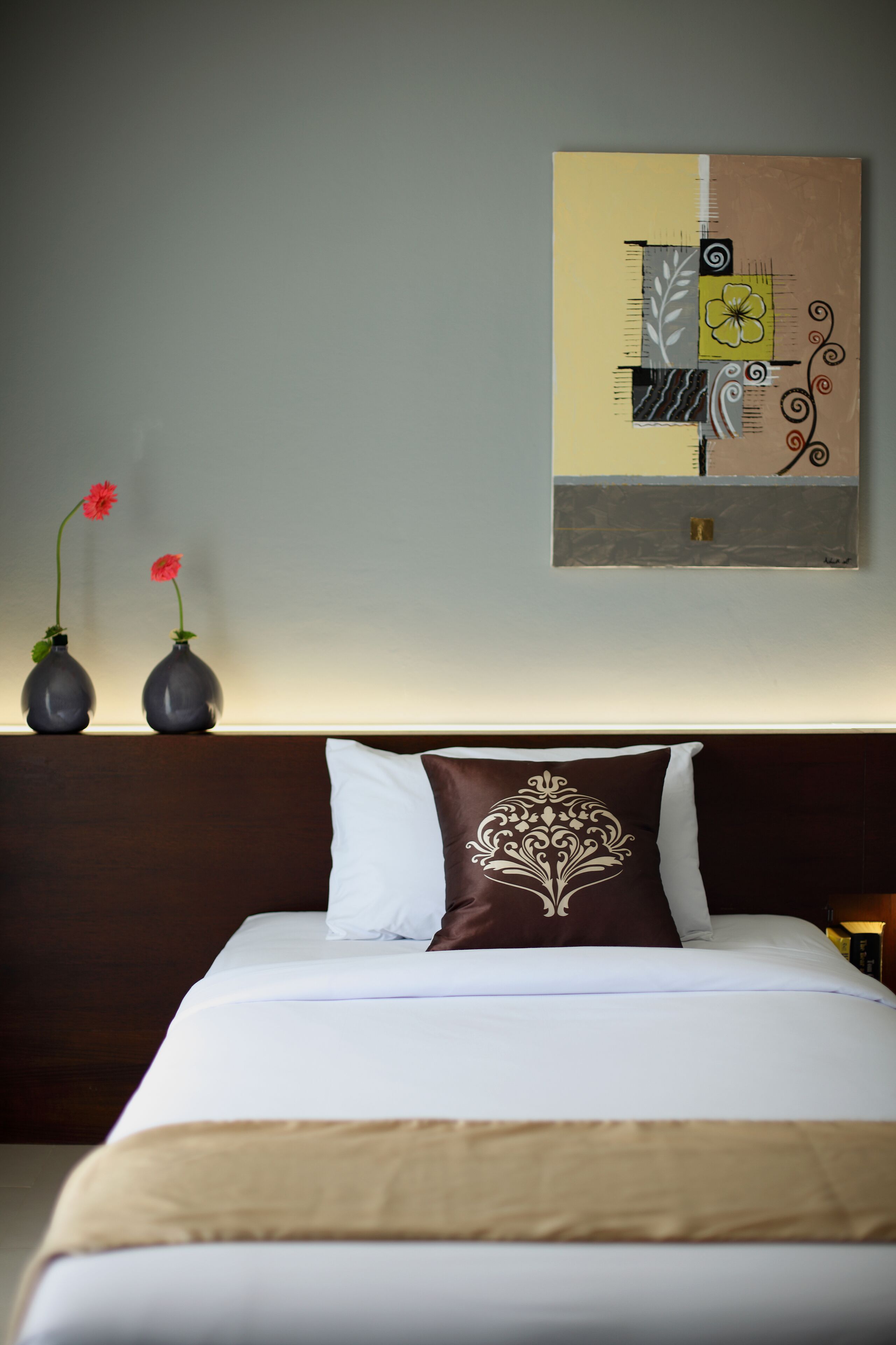 A neatly made bed with a white and brown pillow, two vases with red flowers, and a wall art piece above the bed.