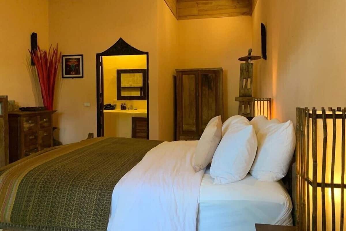 Quarto com cama, cabeceira de madeira, mesa de cabeceira, cômoda, espelho e banheiro visível por uma porta aberta.