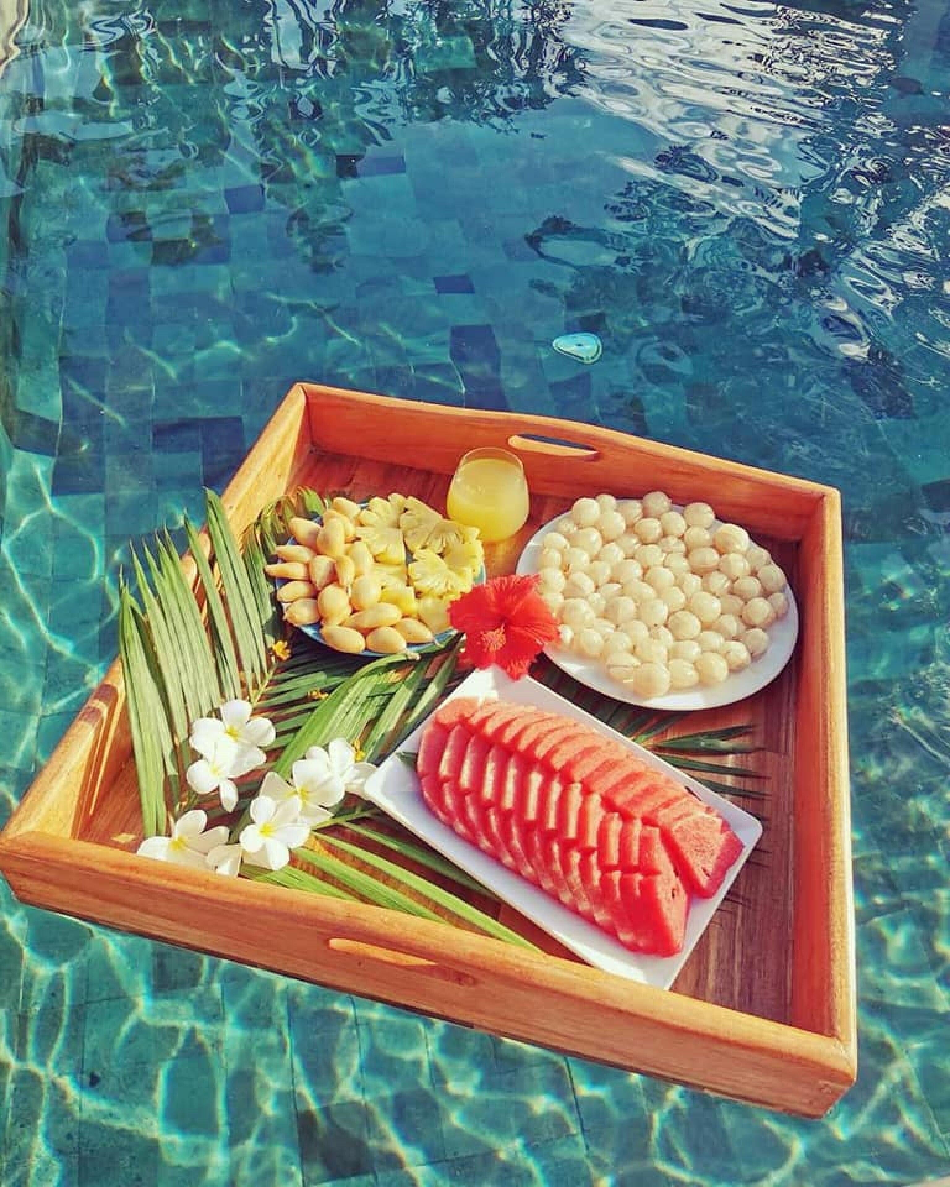 Una bandeja de madera con sandía en rodajas, piña y un tazón con esferas blancas, posiblemente perlas de tapioca, junto a una piscina.