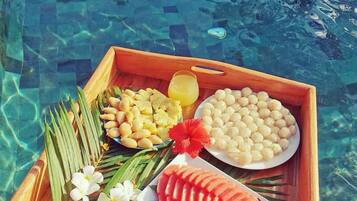 Una bandeja de madera con sandía en rodajas, piña y un tazón con esferas blancas, posiblemente perlas de tapioca, junto a una piscina.