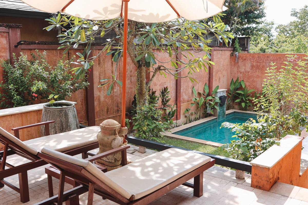 A poolside area with wooden lounge chairs and a stone statue, surrounded by lush greenery and a brick wall.