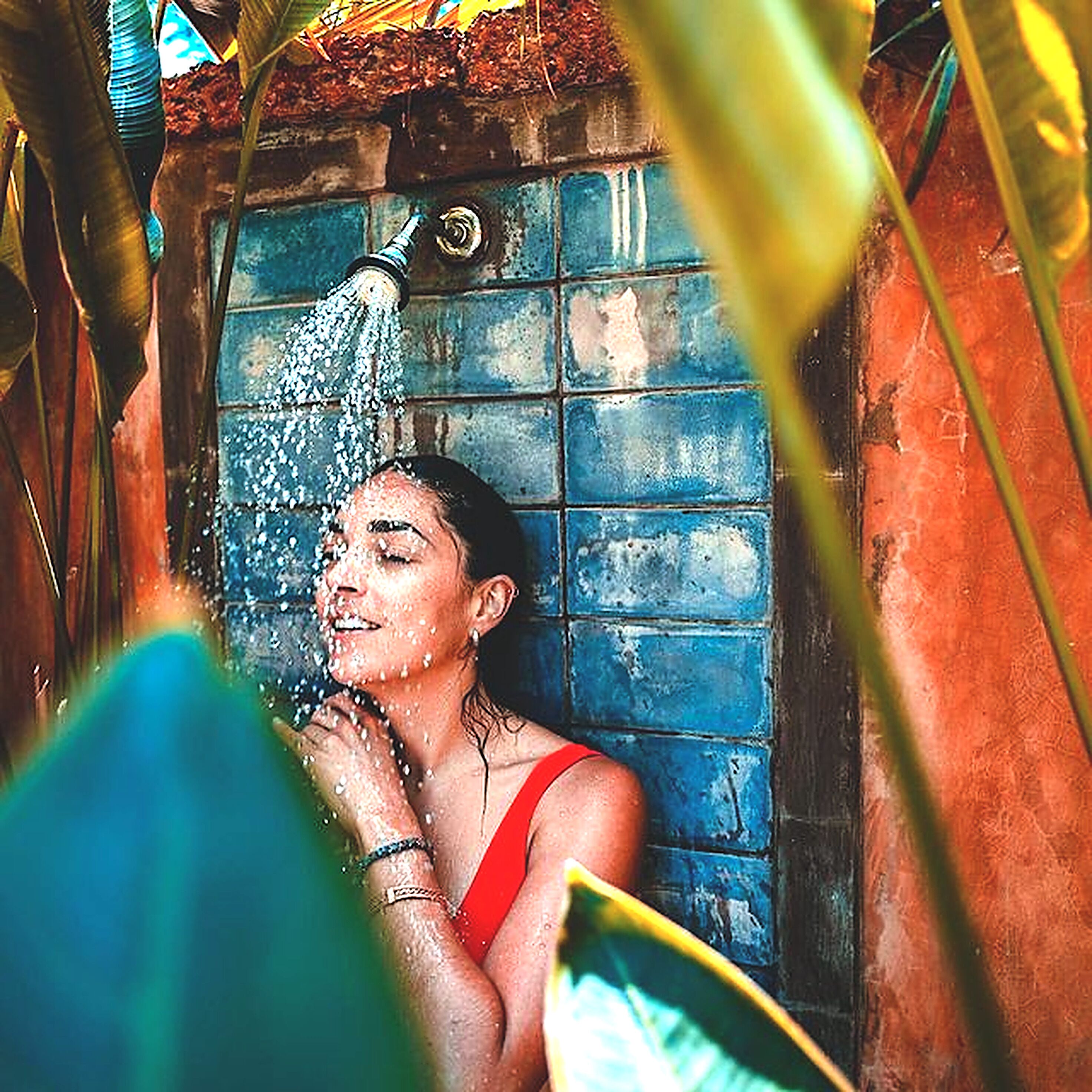 Una persona con remera roja bajo una ducha, con agua corriendo, rodeada por un muro de azulejos azules y follaje.