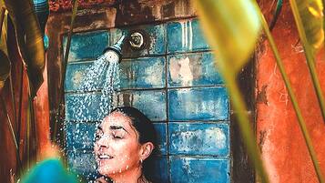 Una persona con remera roja bajo una ducha, con agua corriendo, rodeada por un muro de azulejos azules y follaje.