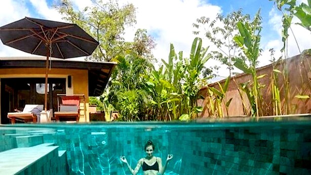A swimming pool with a statue of a woman in the water, surrounded by lush greenery and a building with a covered patio.