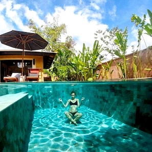A swimming pool with a statue of a woman in the water, surrounded by lush greenery and a building with a covered patio.