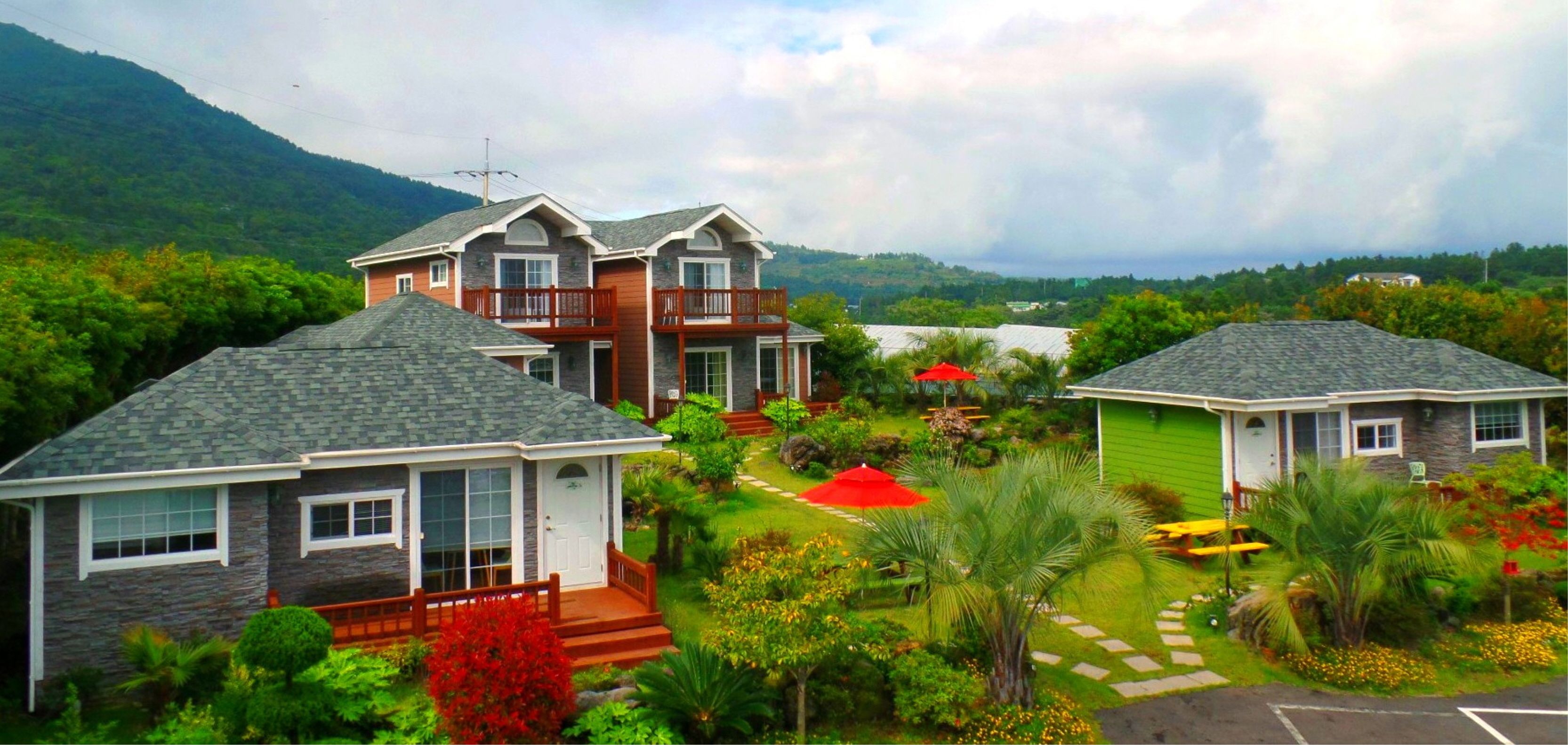 A residential area with two houses, one with a balcony and another with a porch, surrounded by lush greenery and a mountainous backdrop.